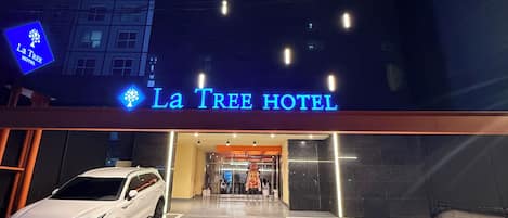 Exterior del Hotel La Tree con una camioneta SUV blanca estacionada frente, una entrada despejada y un edificio con múltiples ventanas.