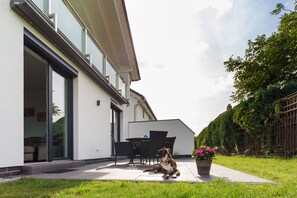 Ein modernes Haus mit einer Terrassenfläche, ein Hund, der auf dem Gras sitzt, und ein Blumentopf mit Blumen.
