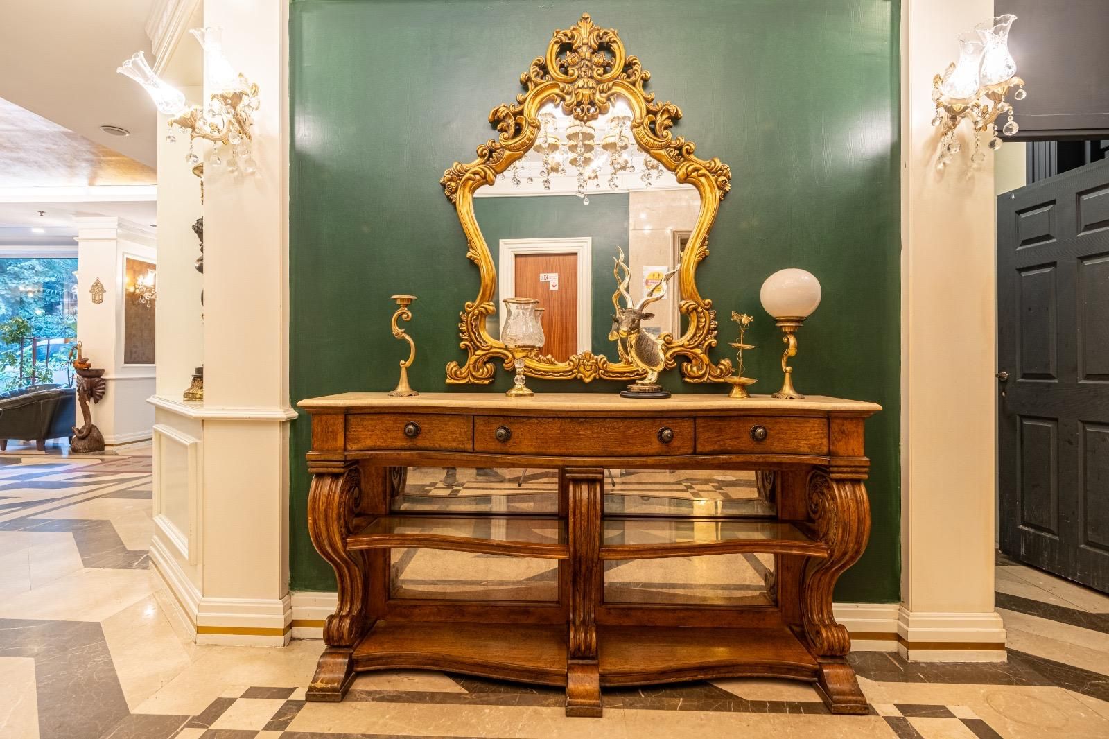 A grand hallway with a large ornate mirror, a wooden console table with drawers, and a chandelier.