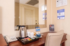 A hotel room with a wooden desk, a telephone, a coffee maker, a tray with snacks, and a mirror reflecting the room.