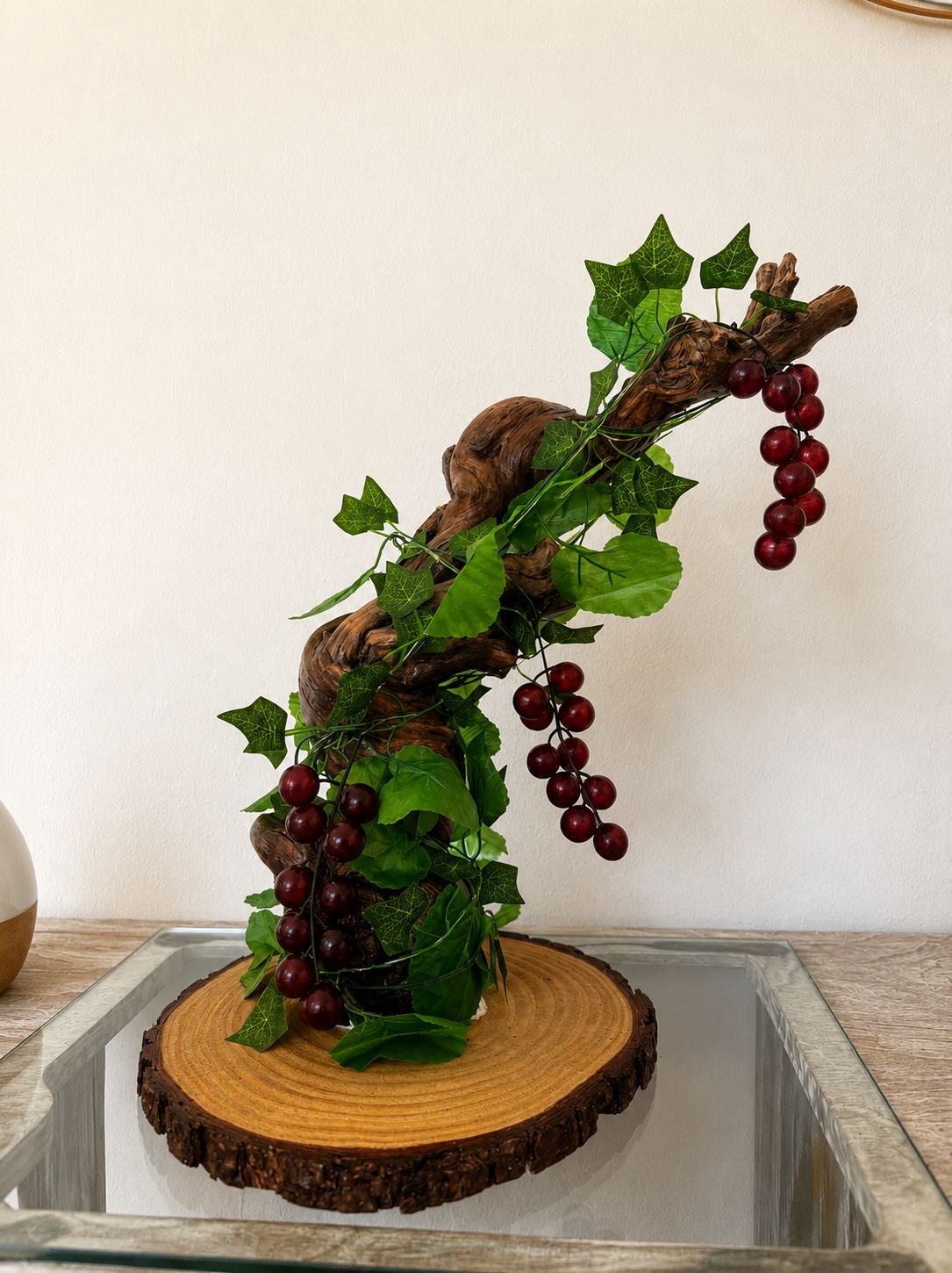 Um ramo de madeira decorativo com folhas verdes e berris vermelhas, sobre uma mesa de vidro com base de madeira.