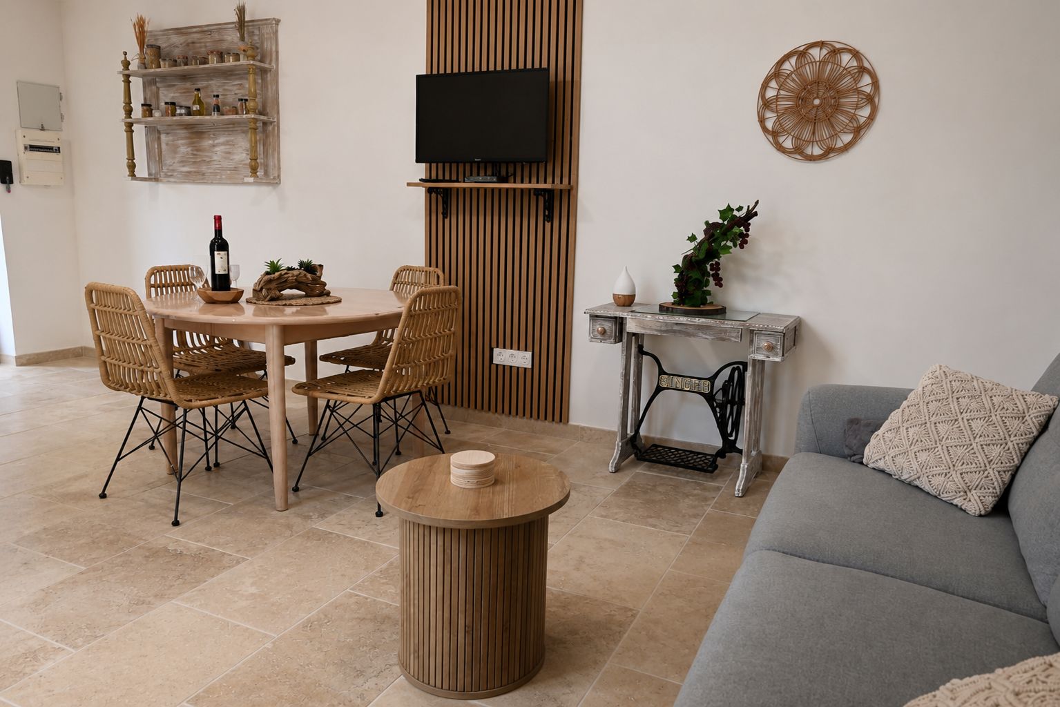 A living room with a round wooden table, wicker chairs, a grey sofa, a television mounted on a wooden panel, and a small table with a plant.