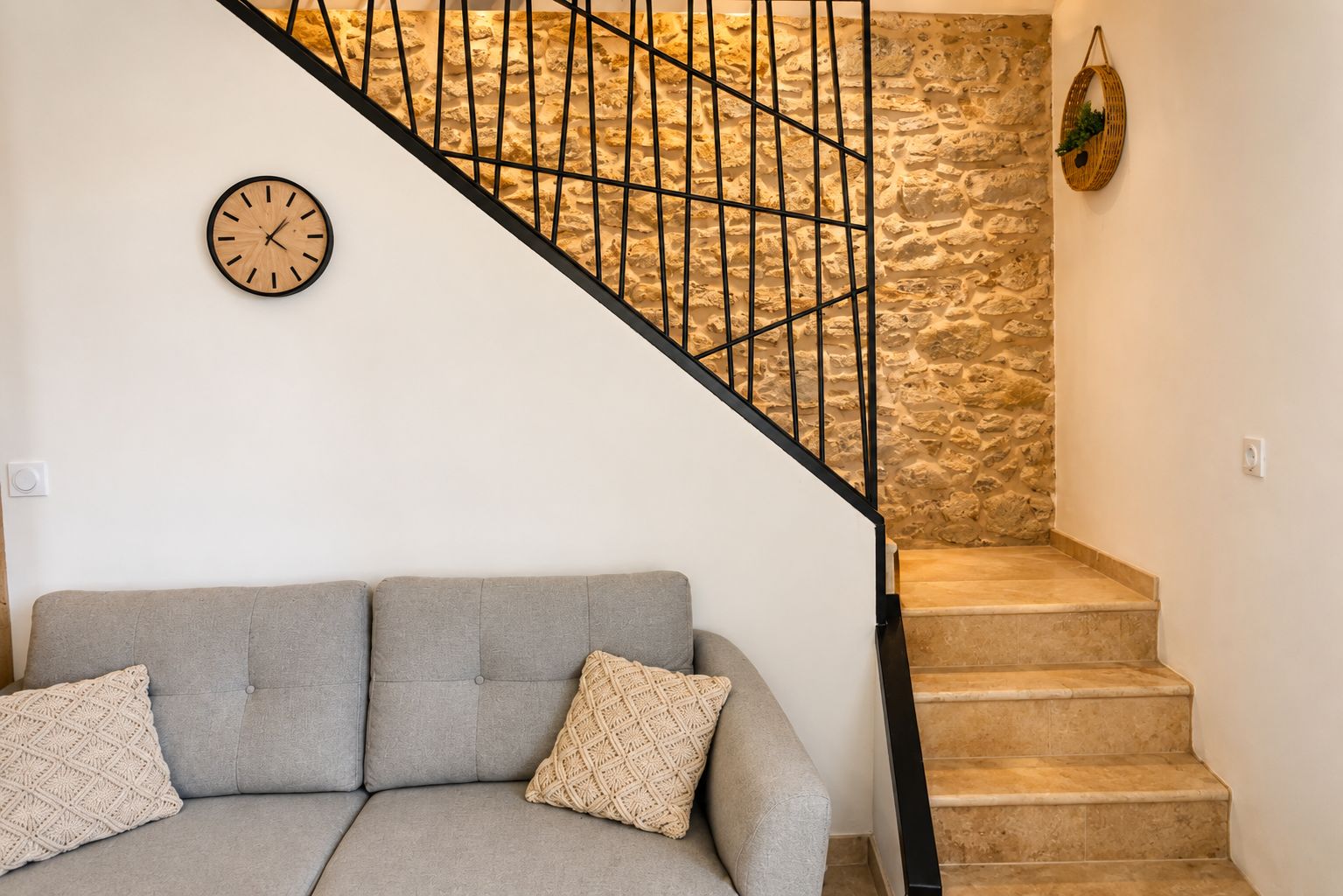 Un escalier avec un mur en pierre, un canapé gris avec des coussins à motifs et une horloge murale.