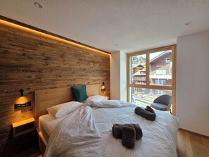 Une chambre d’hôtel moderne avec une tête de lit en bois, un grand lit avec du linge de lit blanc, un oreiller vert et une vue sur un immeuble par la fenêtre.