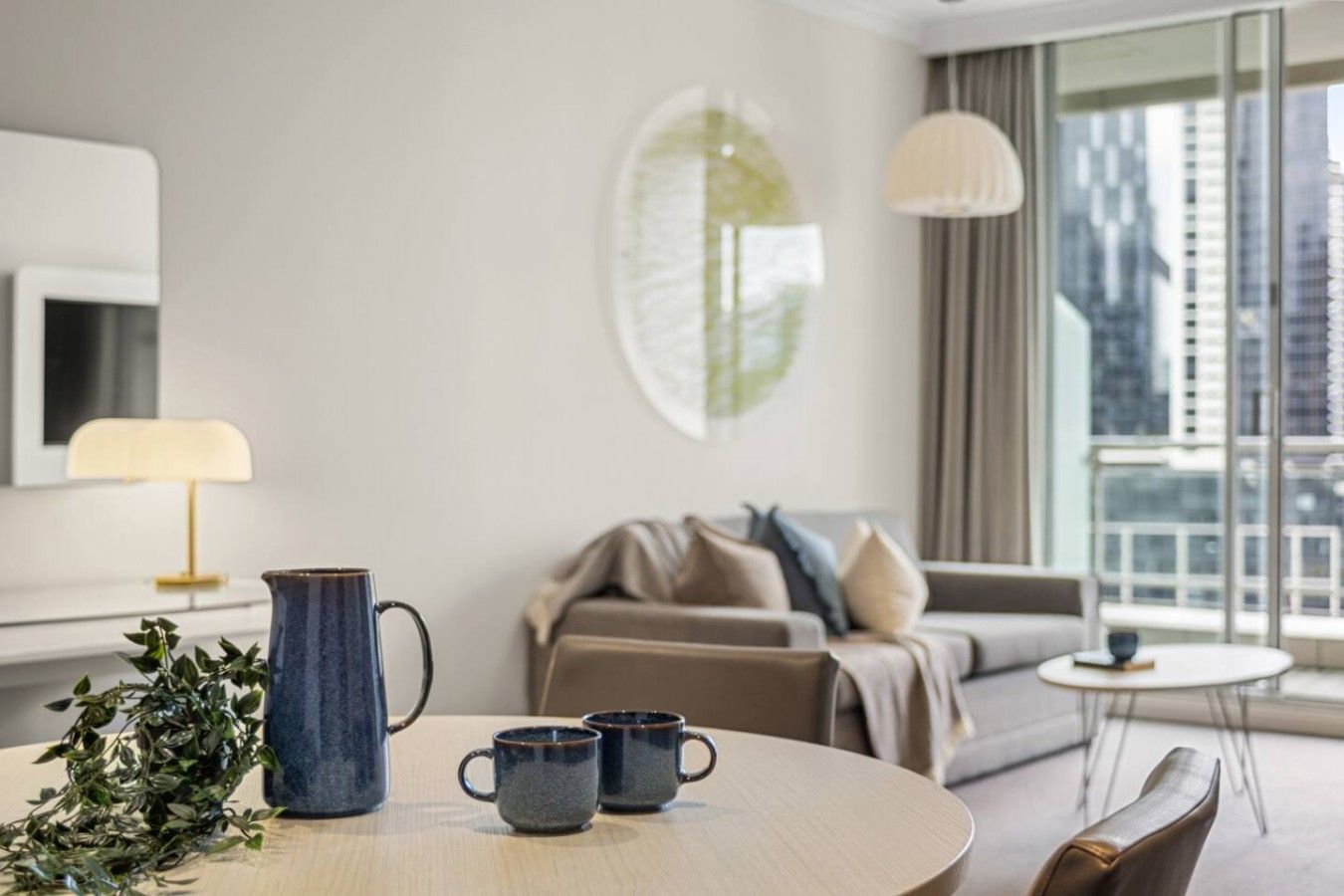 A modern hotel room with a round dining table, two mugs, a pitcher, and a plant.