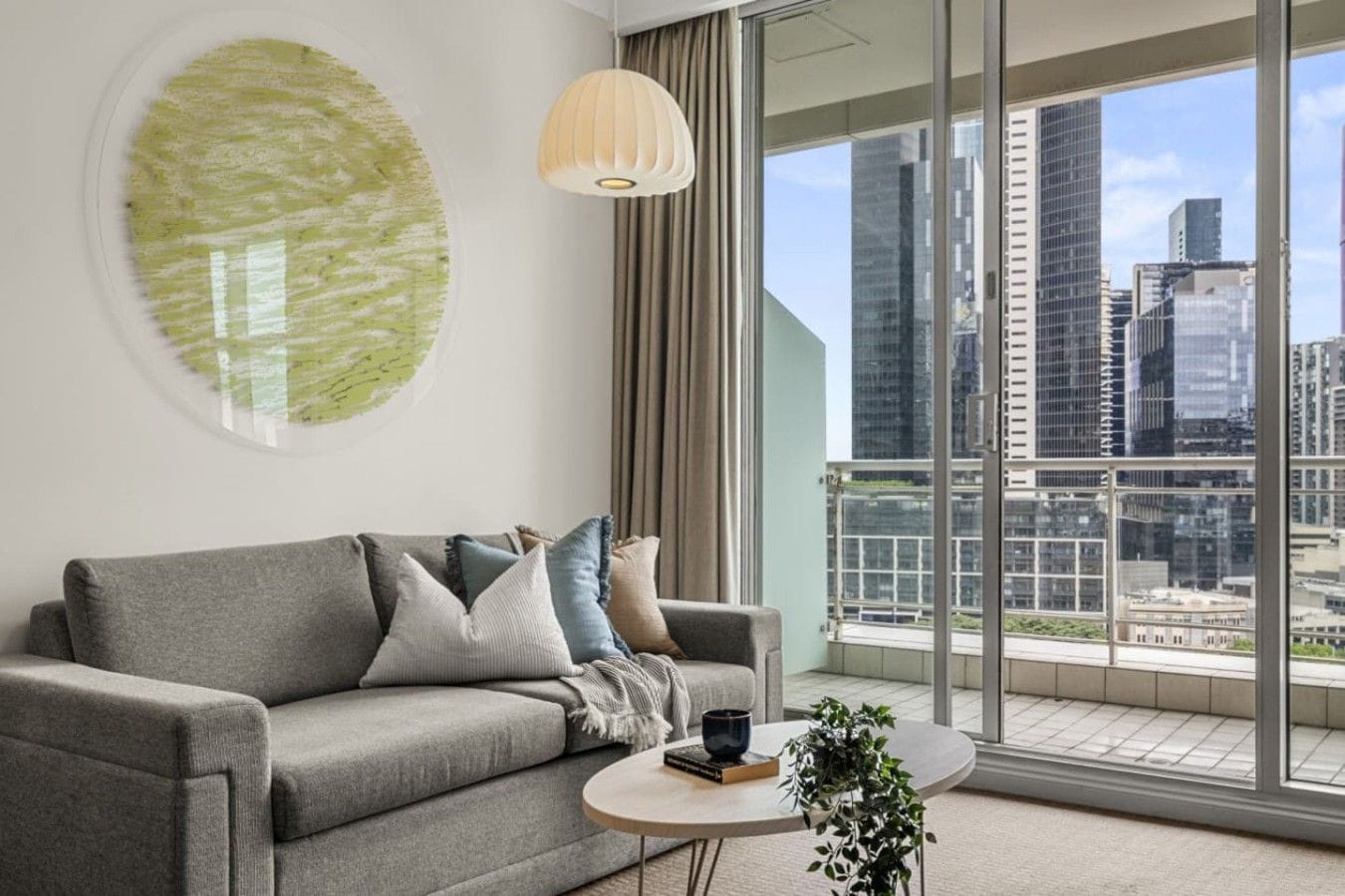 A modern living room with a grey sofa, a round coffee table, and a city view through large windows.