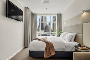Camera d'albergo con un letto grande, una TV a schermo piatto fissata al muro, una finestra con vista sulla città e un comodino con un libro e una tazza.