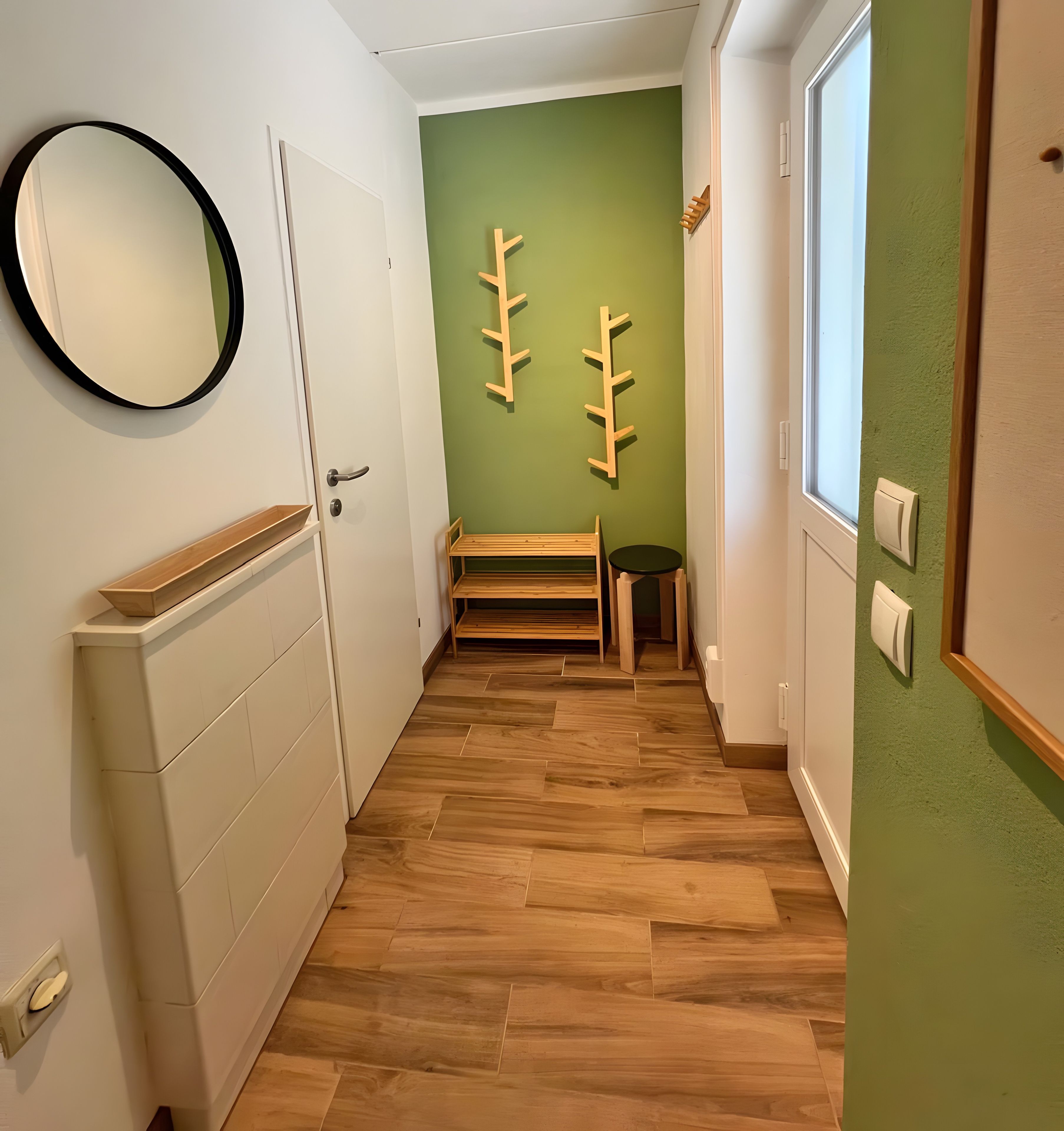 A hallway with a wooden floor, a white door, a mirror, and a green wall with a wooden rack.
