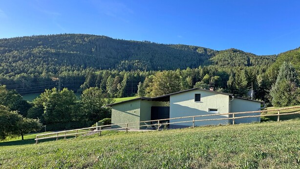 A small building with a flat roof, surrounded by a wooden fence, set against a backdrop of a forested mountain.