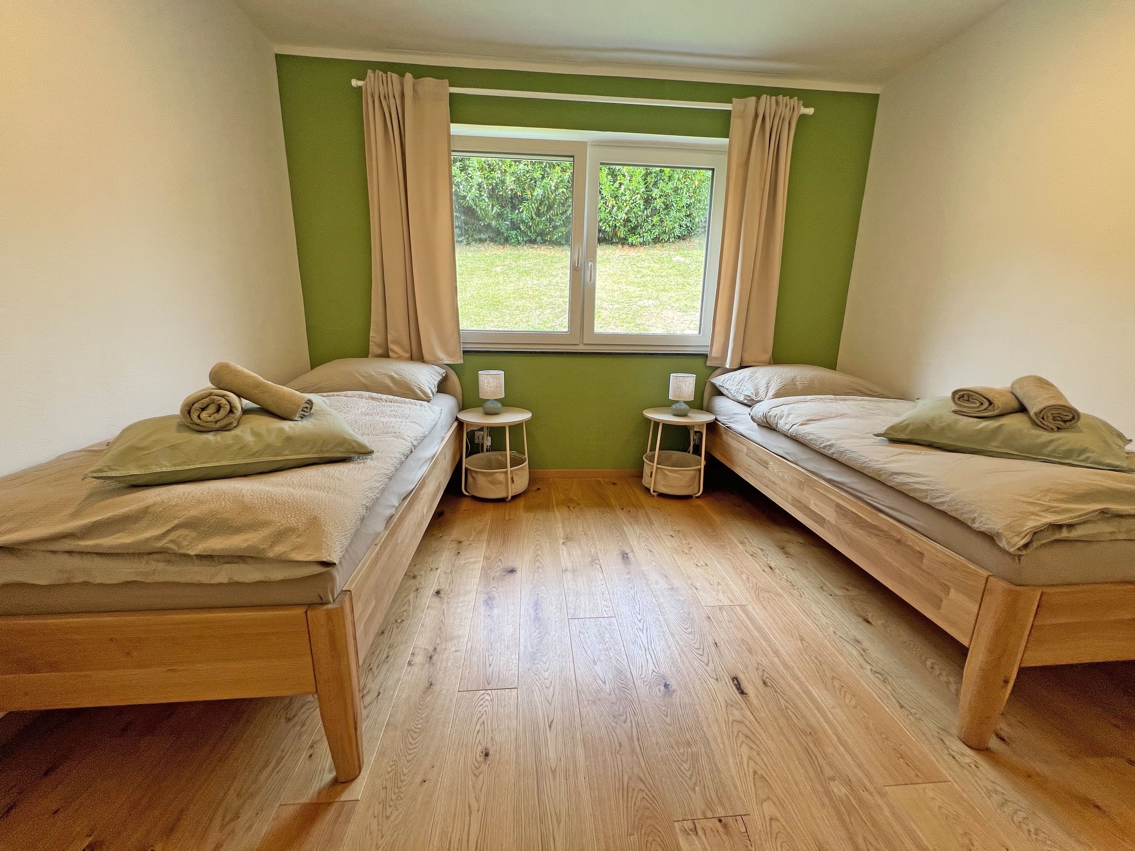 Une chambre avec deux lits simples, une fenêtre avec des rideaux, une petite table avec une lampe, et une vue sur la verdure à l’extérieur.