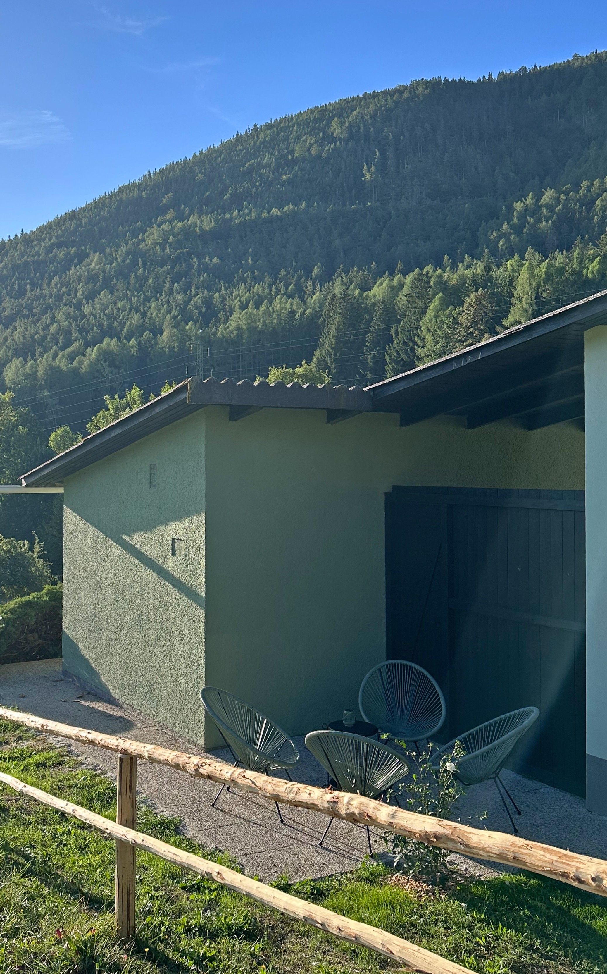 Ein kleines Gebäude mit einem geneigten Dach, einer gepflasterten Terrasse mit Metallstühlen und einem bewaldeten Hügel im Hintergrund.