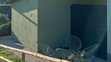 Ein kleines Gebäude mit einem geneigten Dach, einer gepflasterten Terrasse mit Metallstühlen und einem bewaldeten Hügel im Hintergrund.