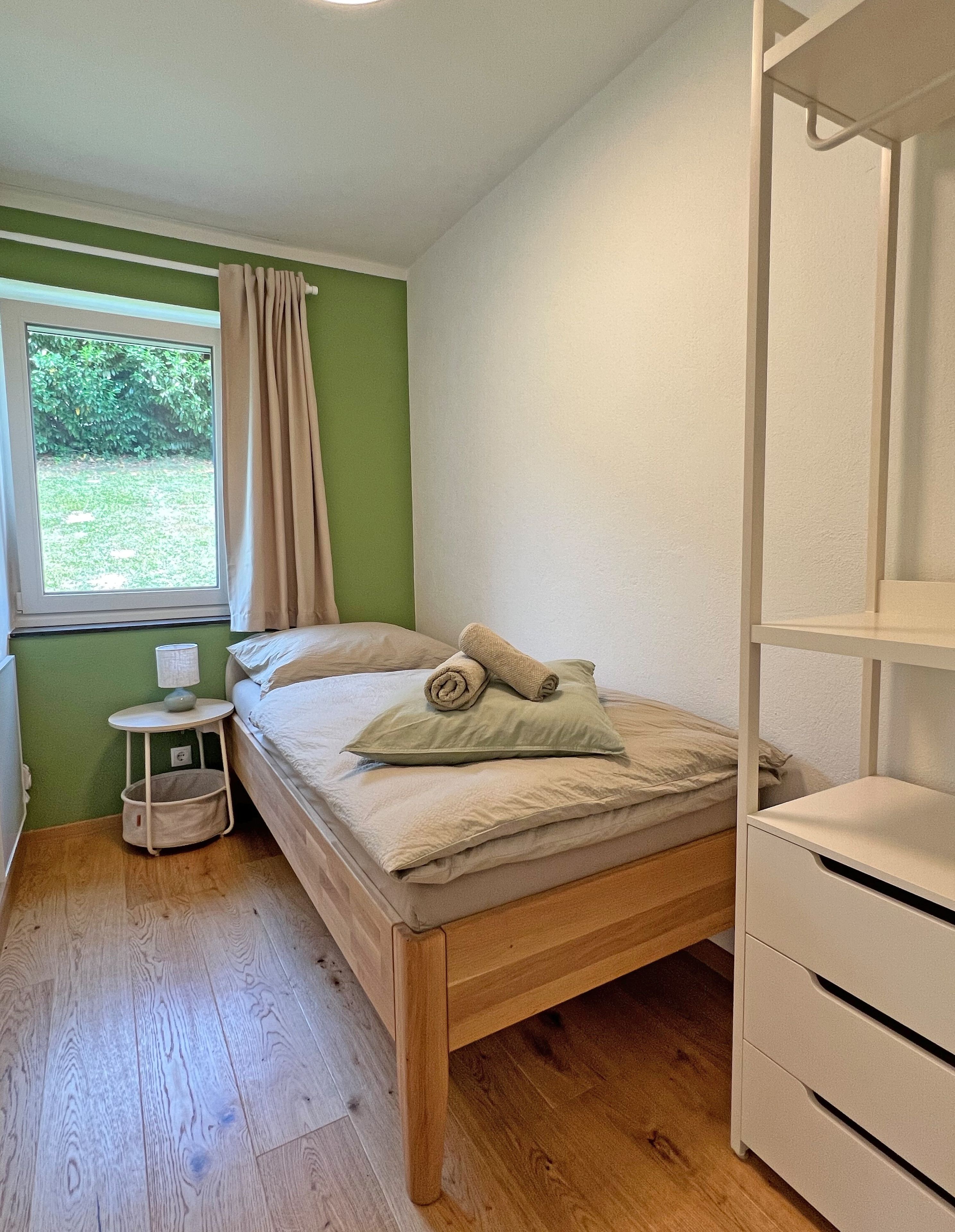 Ein Einzelbett mit Holzrahmen, ein kleiner Nachttisch mit Lampe und ein Fenster mit Vorhängen.