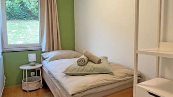Ein Einzelbett mit Holzrahmen, ein kleiner Nachttisch mit Lampe und ein Fenster mit Vorhängen.