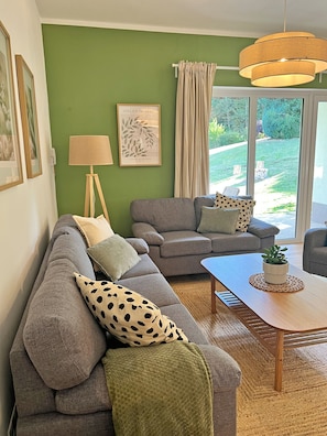 A living room with a sectional sofa, a coffee table, and a lamp. There is a window with curtains and a view of the outdoors.