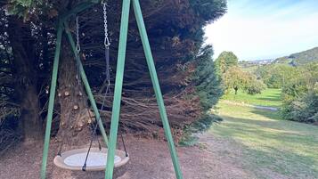 Eine Schaukel unter einem großen Baum in einer grünen Wiese mit Blick auf eine entfernte Landschaft.