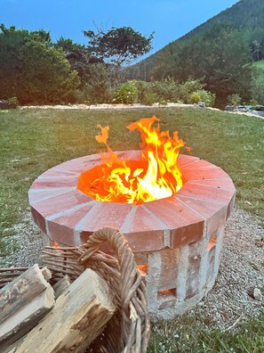 Ein rundes Feuerbecken mit Flammen, umgeben von einer Wiese und einem Berg im Hintergrund.