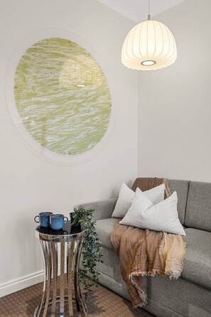 A room with a grey sofa, a round wall decoration, a hanging light fixture, a small side table with a plant, and two blue cups. - Central City Studio: King Bed + Amazing Views! (Melbourne)