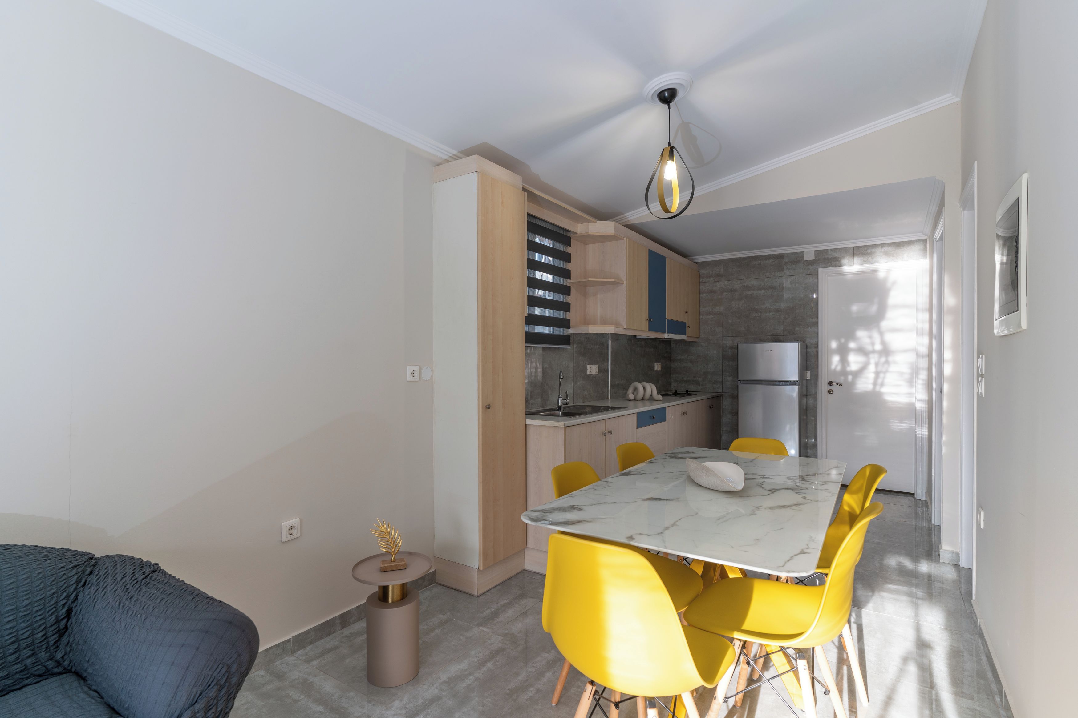 Cozinha compacta com eletrodomésticos modernos, área de jantar com cadeiras amarelas e um sofá cinza.