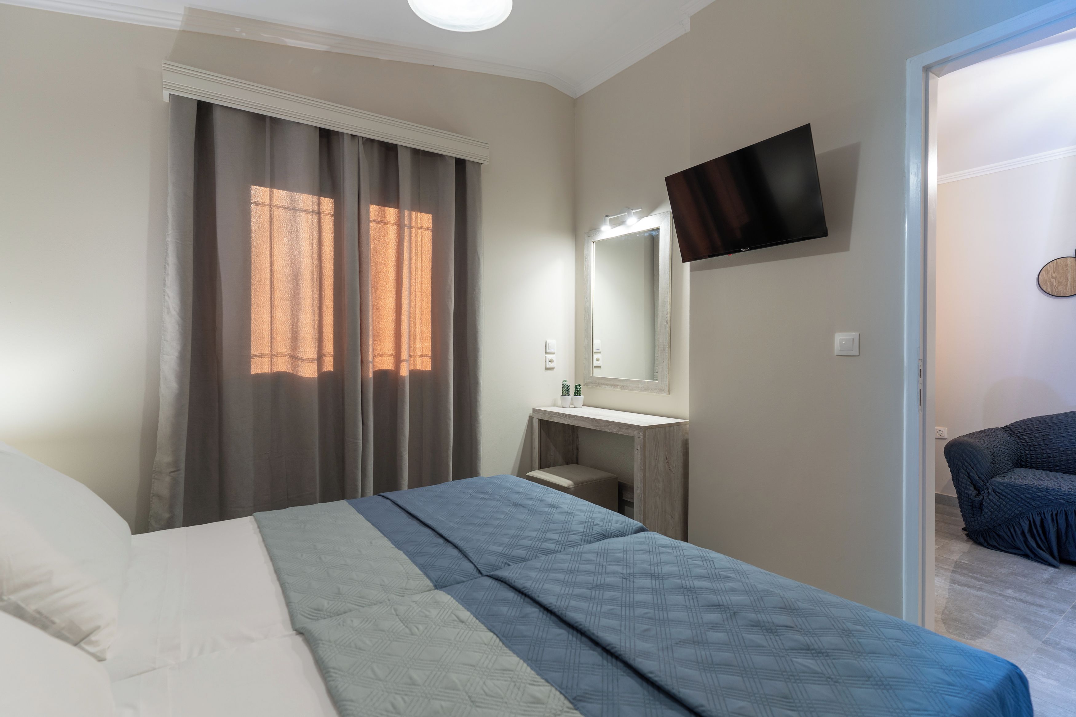 Quarto com cama, mesa de cabeceira, espelho, TV fixada na parede e porta que dá para outro espaço.