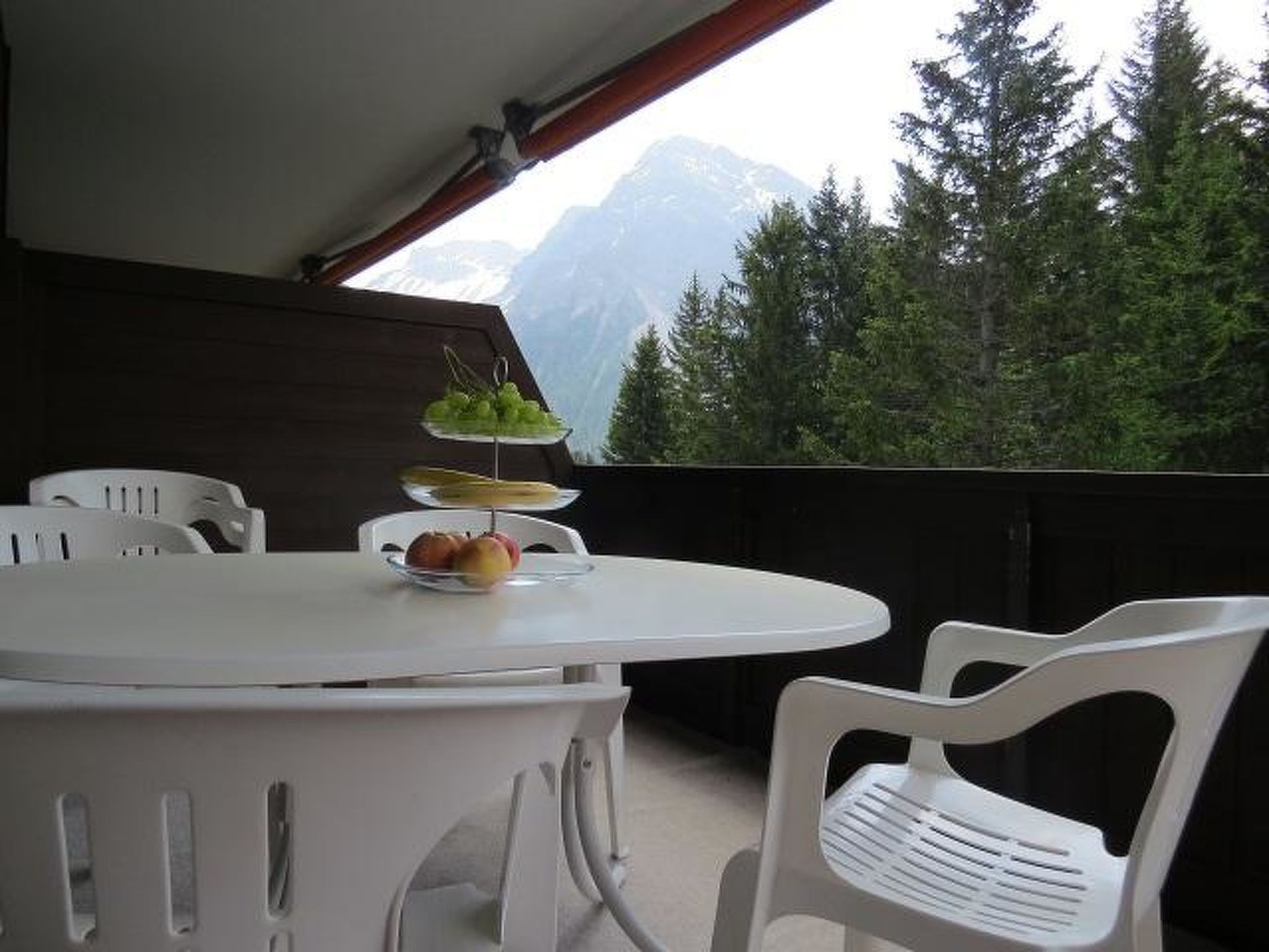 A balcony with a white table and chairs, a plate of fruit, and a view of a forest and mountains.