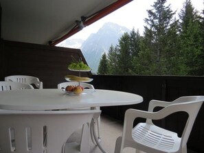 Un balcón con una mesa y sillas blancas, una fuente de fruta y vistas a un bosque y montañas.