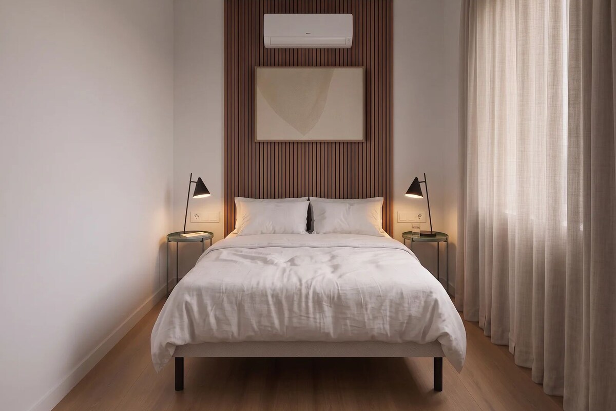 A modern bedroom with a bed, two bedside tables with lamps, a wall-mounted air conditioner, and a large window with curtains.