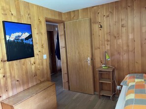 Ein Zimmer mit Holzwandverkleidung, einem Bett mit bunter Decke, einem Schreibtisch mit Lampe und einem Fernseher, der eine Berglandschaft zeigt.