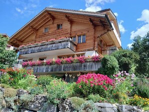 A wooden chalet with a balcony adorned with flowers, surrounded by a garden with various plants and flowers. - Haus Abäsonne Appartement/Fewo, Bad, WC, 1 Schlafraum 2.5 Zimmerwohnung by Interhome (Adelboden)