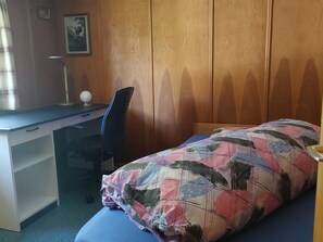 Ein Einzelbett mit einer farbenfrohen Steude, ein Schreibtisch mit Stuhl und eine Lampe.