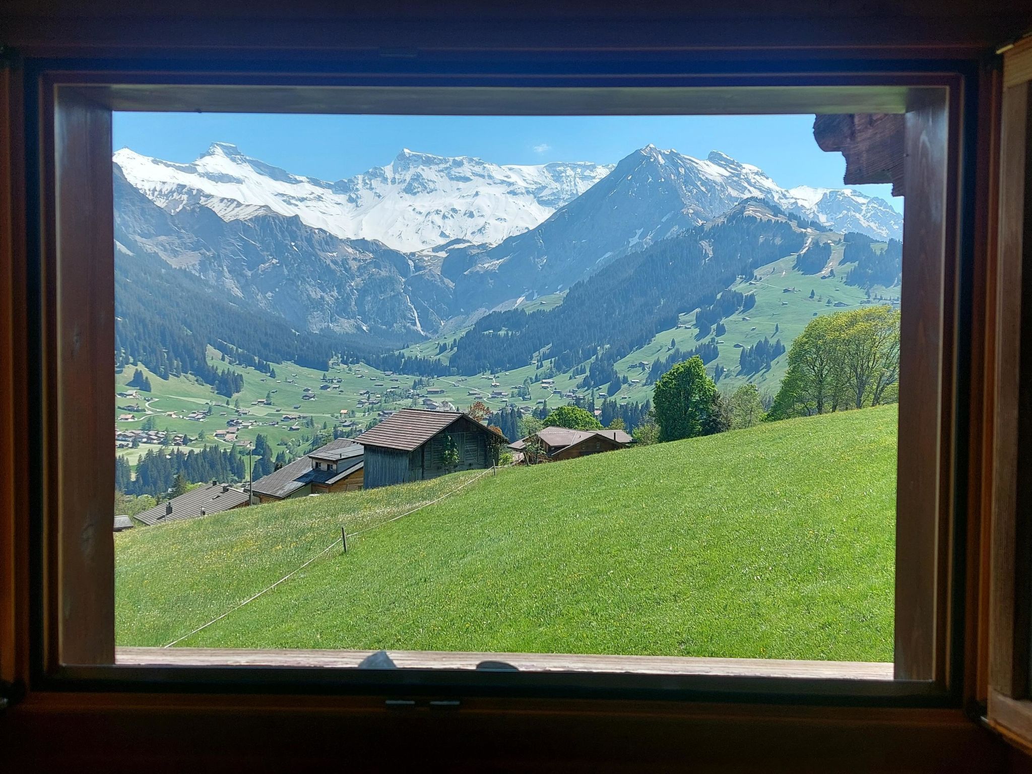 A window view of a mountainous landscape with snow-capped peaks, a green valley, and traditional wooden houses.