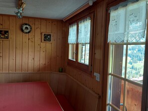 Ein Zimmer mit Holzwänden, einem roten Tisch, zwei Fenstern mit Spitzenvorhängen und einer Uhr an der Wand.