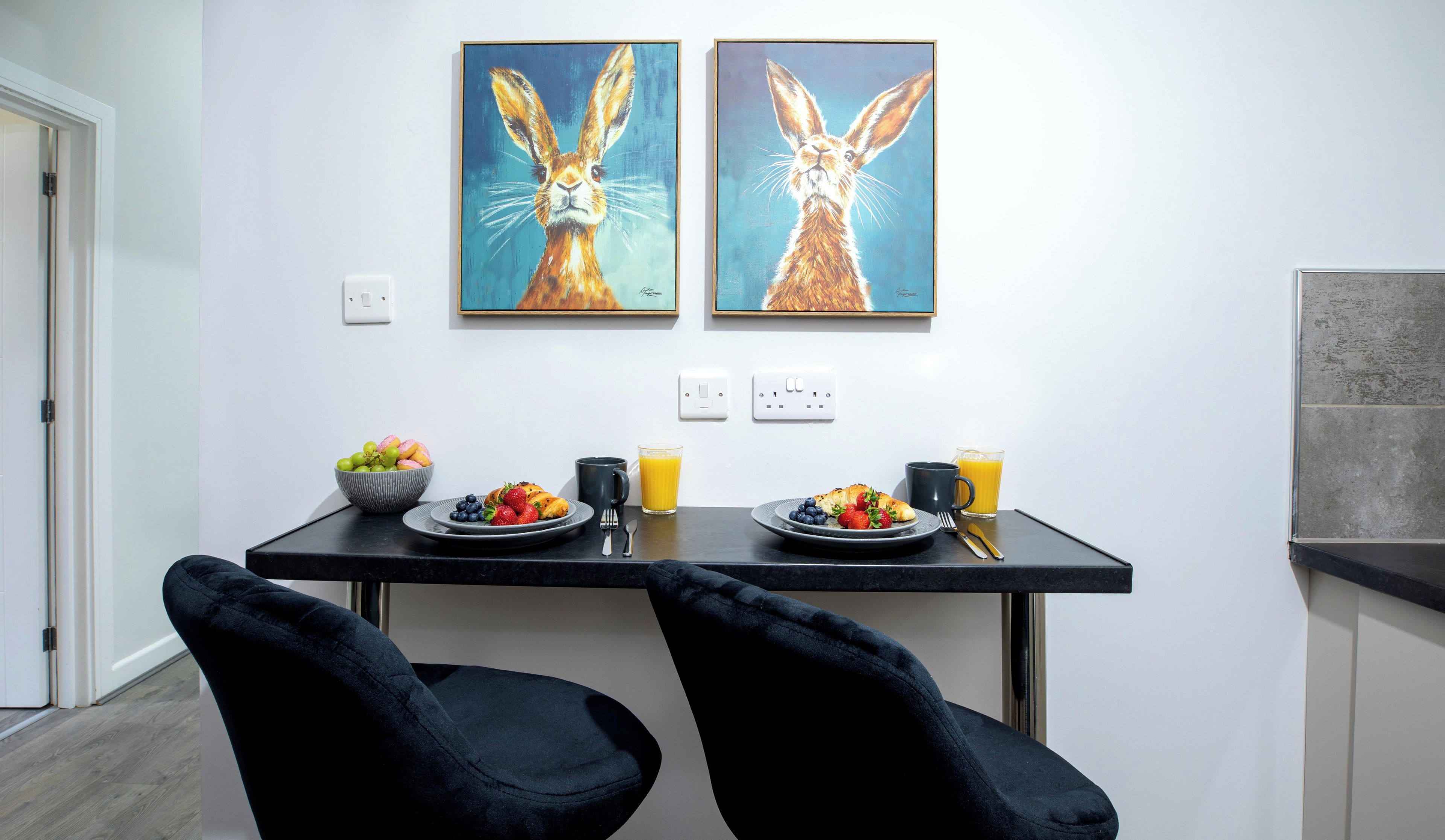 A dining area with two paintings of rabbits, a table with breakfast, and black chairs.