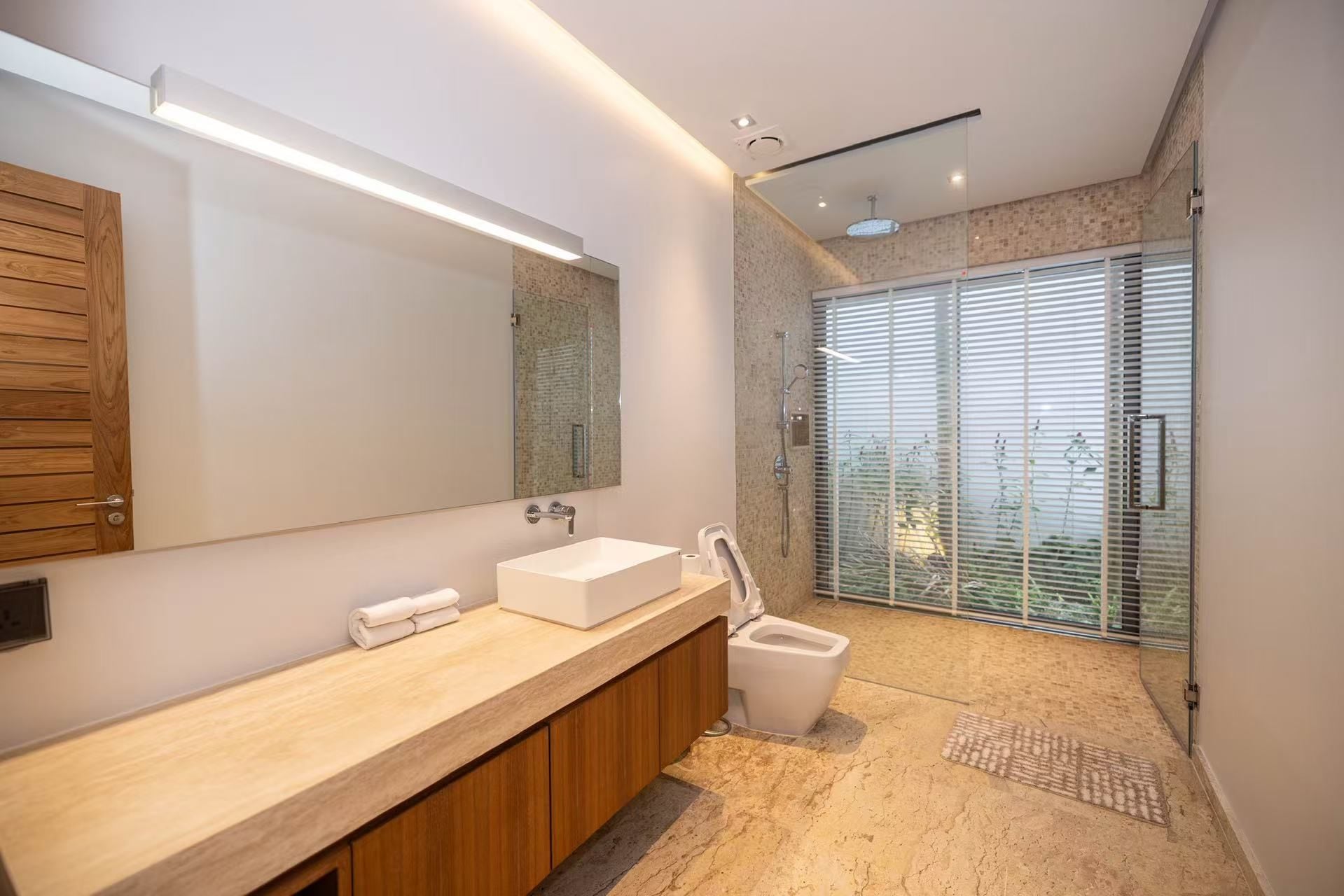 A modern bathroom with a large mirror, a wooden vanity, a shower, and a glass door leading to a balcony.