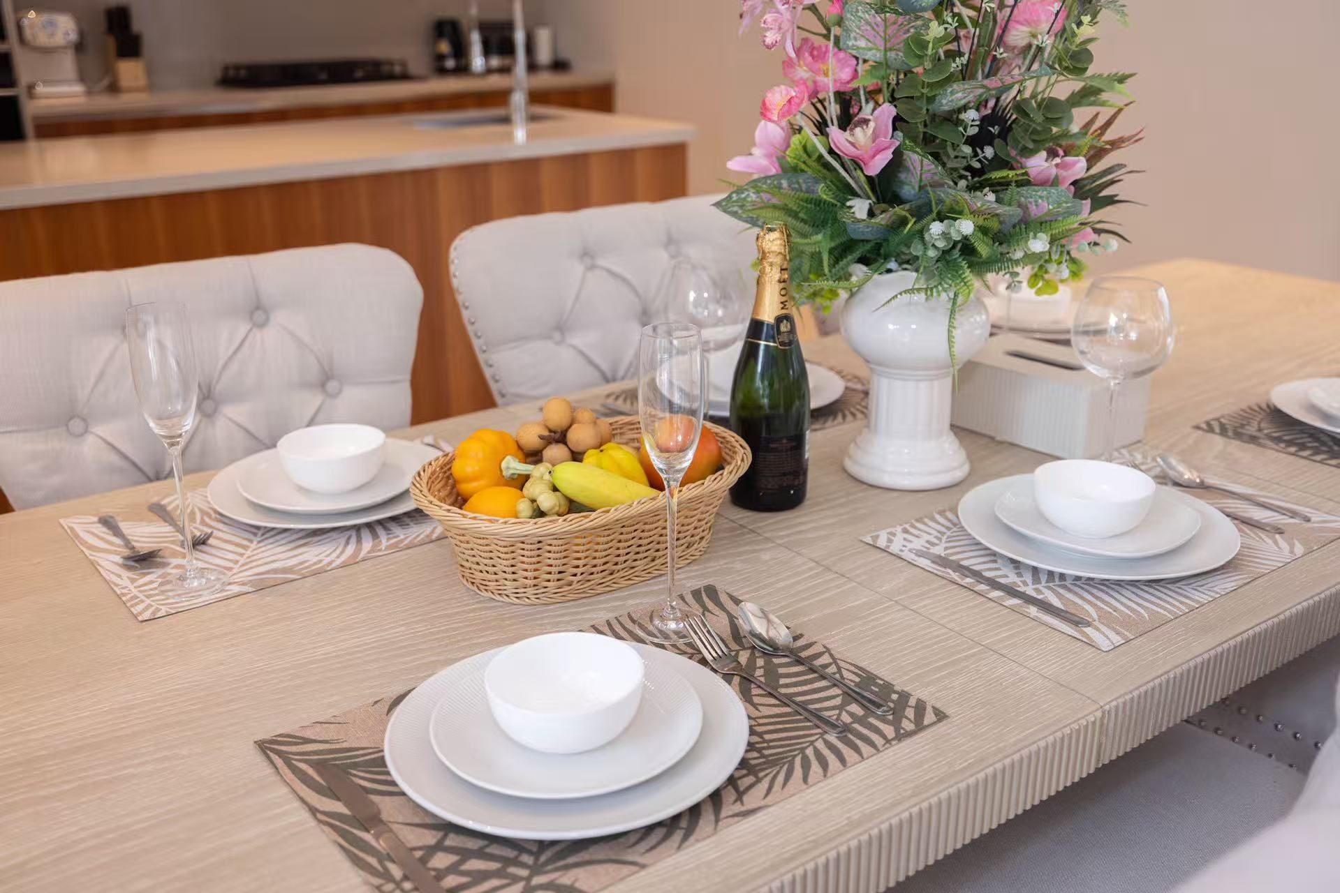 Une table à manger dressée avec des assiettes, des bols, un panier de fruits, une bouteille de champagne et un vase de fleurs.