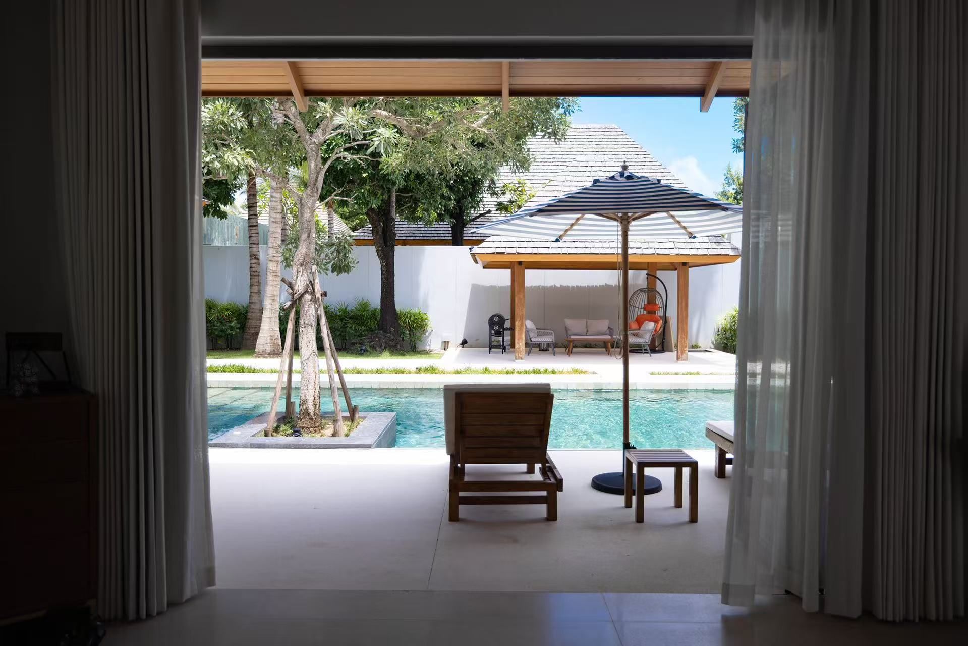 A view of a pool area with a wooden deck, lounge chairs, and a covered seating area with a striped umbrella.