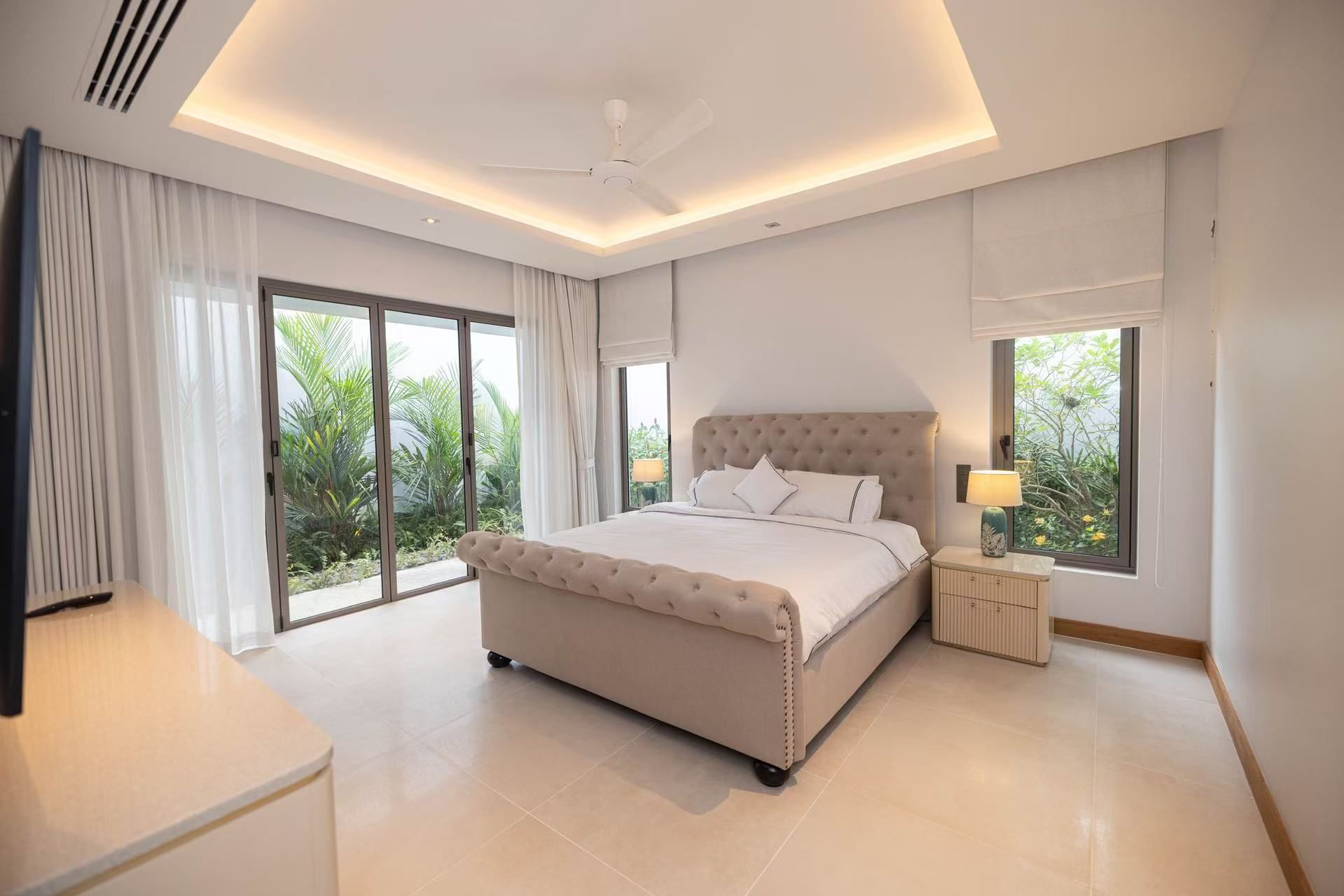 A modern bedroom with a large bed, a ceiling fan, and a view of greenery outside.