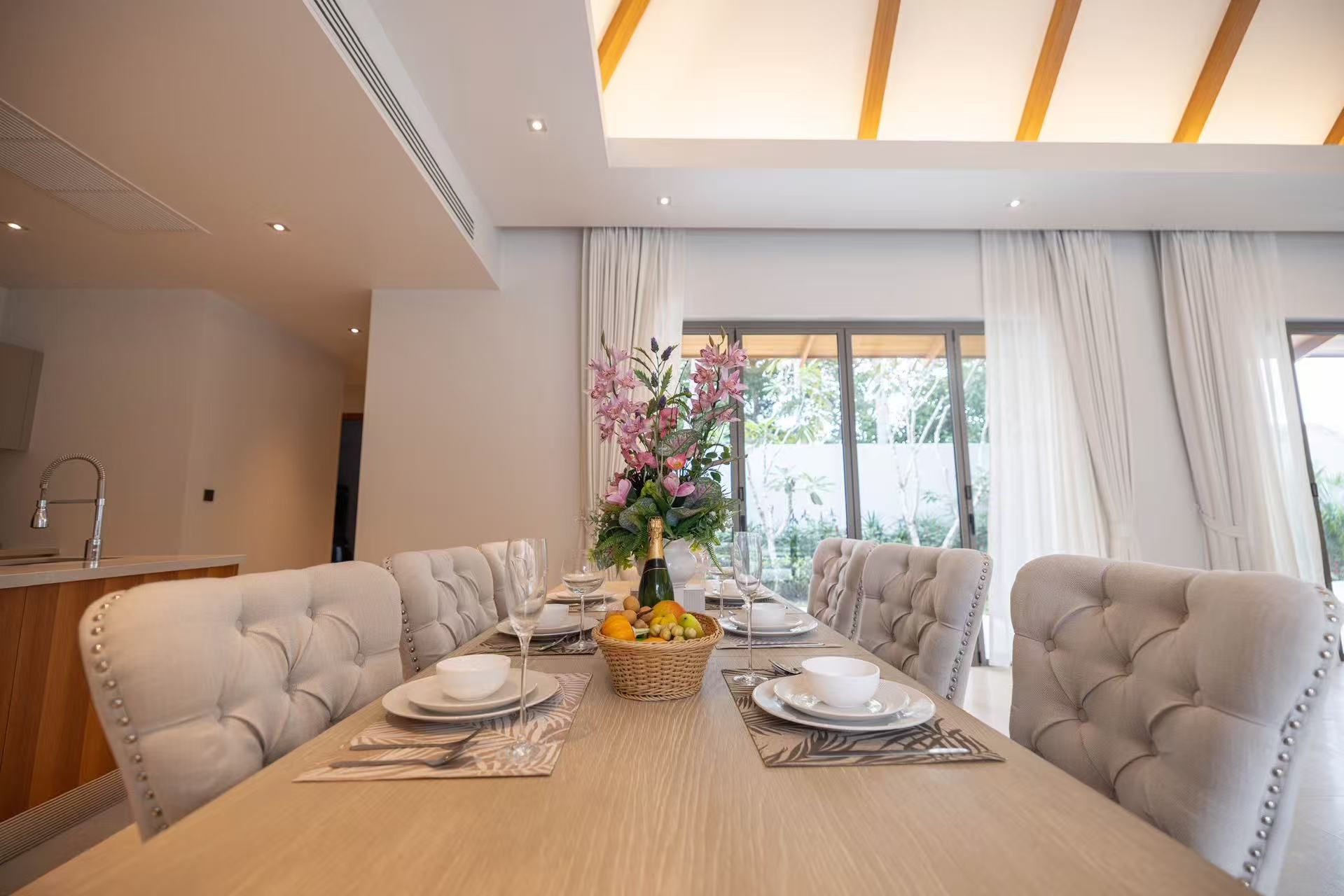 A dining area with a long table set for four, surrounded by upholstered chairs, a vase with flowers, and a fruit basket.