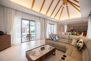 A modern living room with a sofa, a coffee table, and a view of a pool and outdoor seating area. - Anchan Bang Tao Beach 4 Bedroom  Villa (Thalang District)