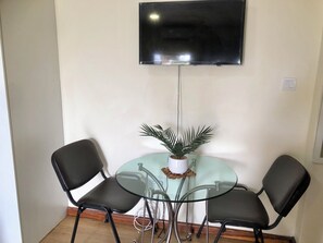 Ein kleines Zimmer mit einem runden Glastisch, zwei schwarzen Stühlen, einer Topfpflanze und einem fest montierten Flachbildfernseher.