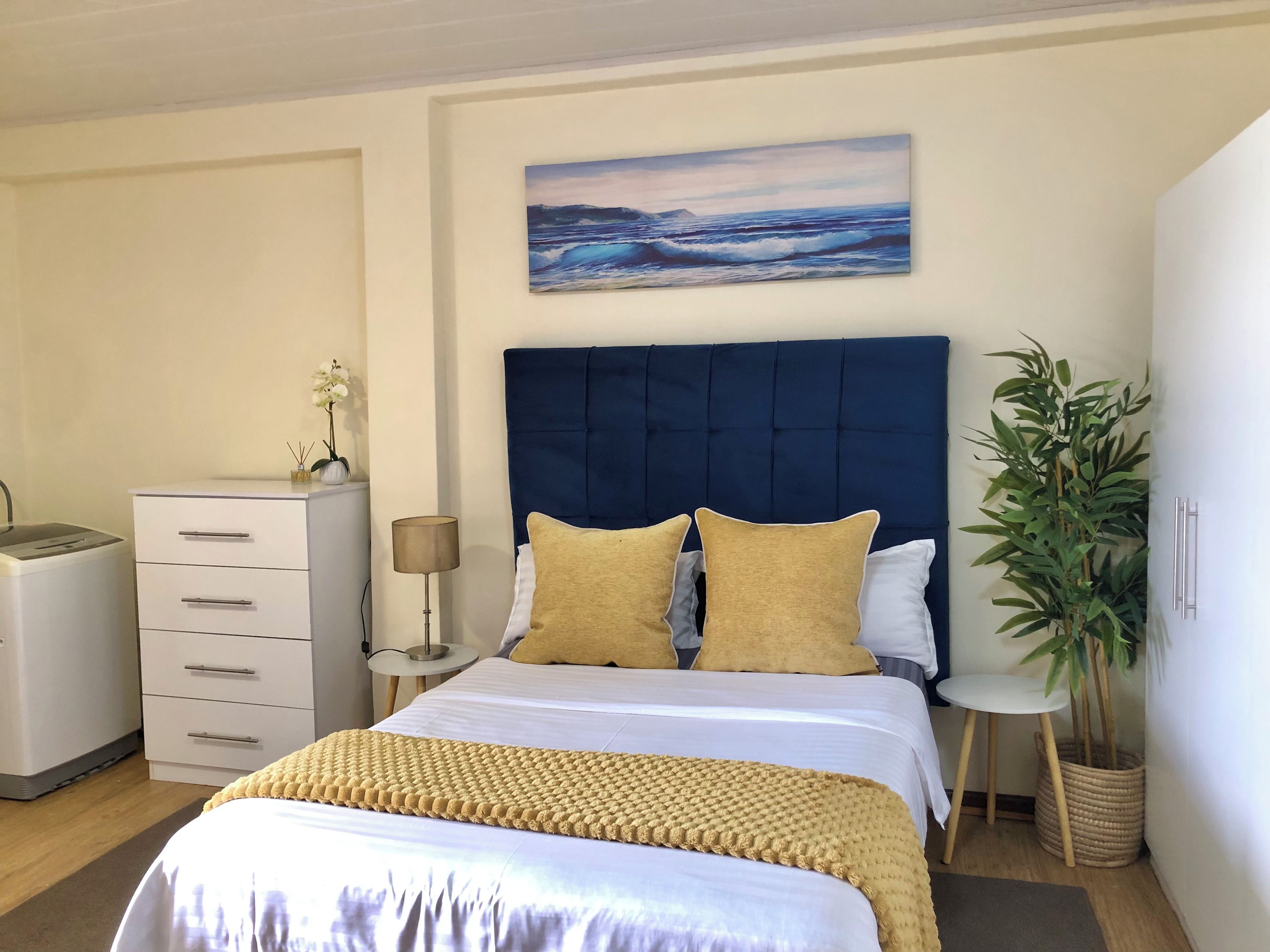 A bedroom with a bed, bedside tables, a lamp, a dresser, a potted plant, and a painting on the wall.
