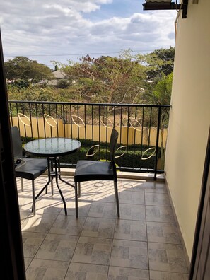 Ein Balkon mit rundem Tisch und zwei Stühlen, einem Fliesenboden und Blick auf Bäume und Gebäude im Hintergrund.