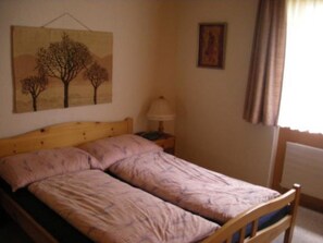 Ein Schlafzimmer mit einem hölzernen Bett, einem Nachttisch mit Lampe, einem Gemälde an der Wand und einem Fenster mit Vorhängen.