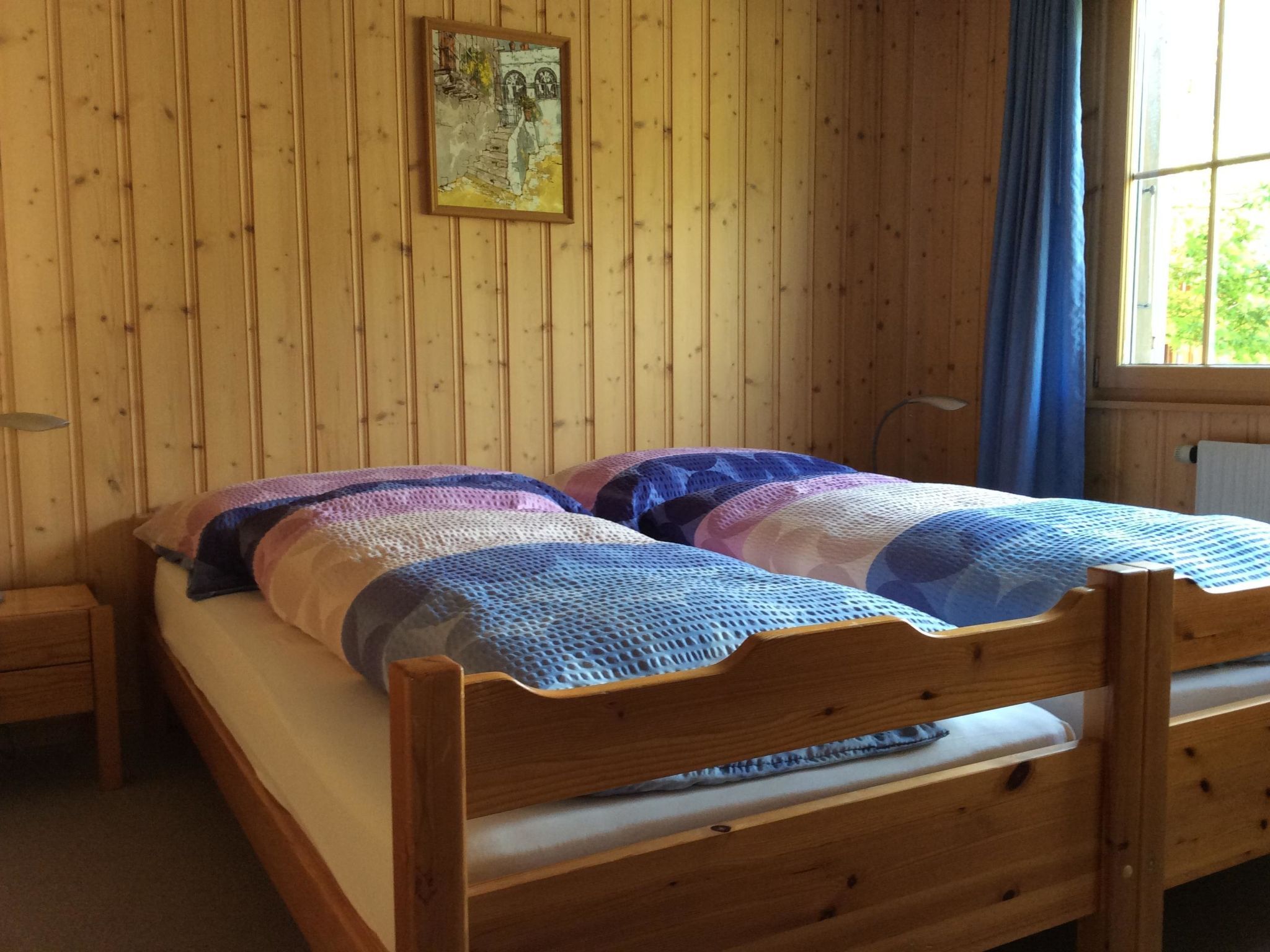 Puisesta sängystä, jossa on sinivalkoinen peitto, yöpöytä, maalauksella koristeltu puuseinä ja sinisellä verholla varustettu ikkuna.