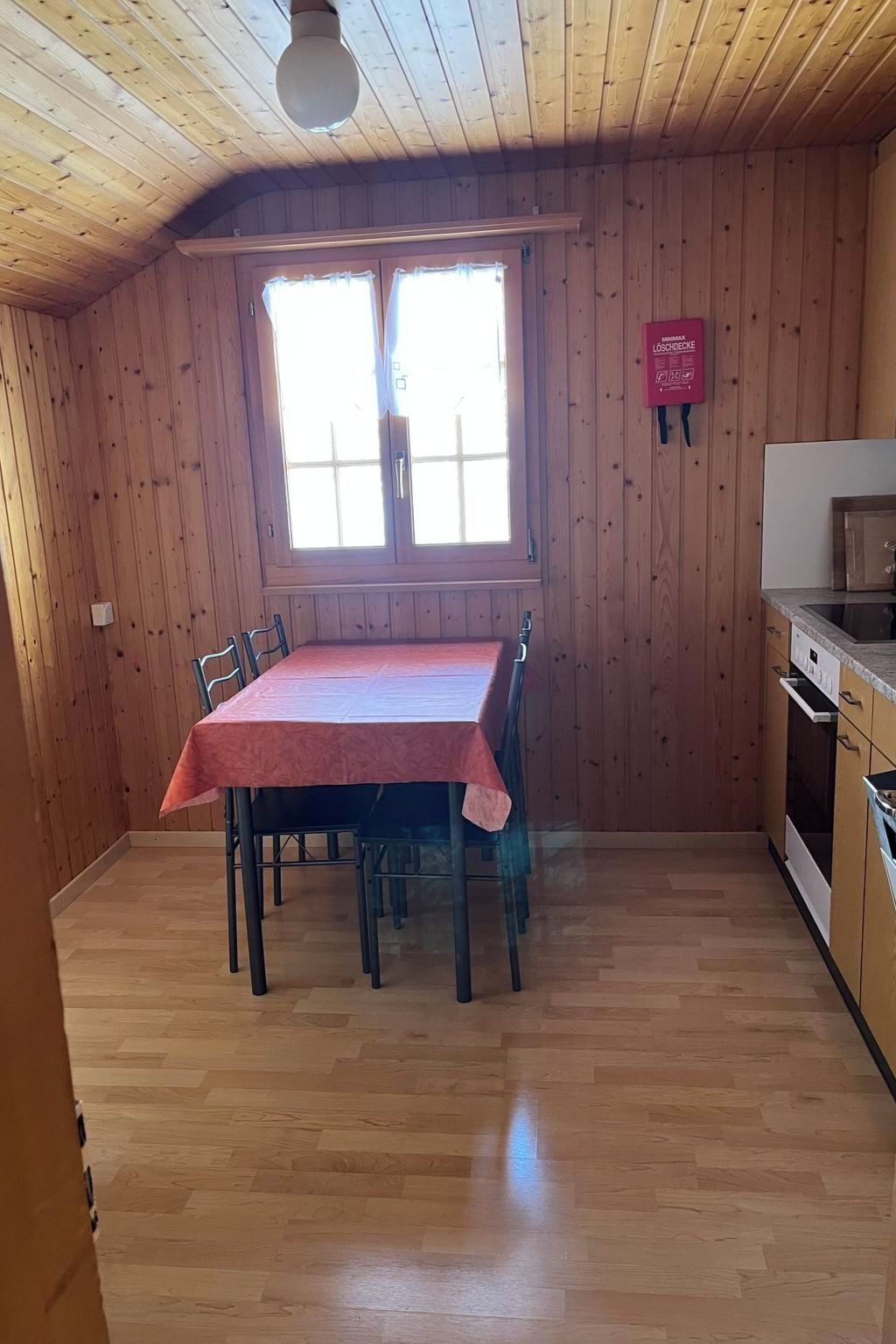Ein Zimmer mit Holzvertäfelung, einem Esstisch mit Stühlen, einem Fenster mit Vorhängen und einer Küchenzeile mit Schränken und Geräten.