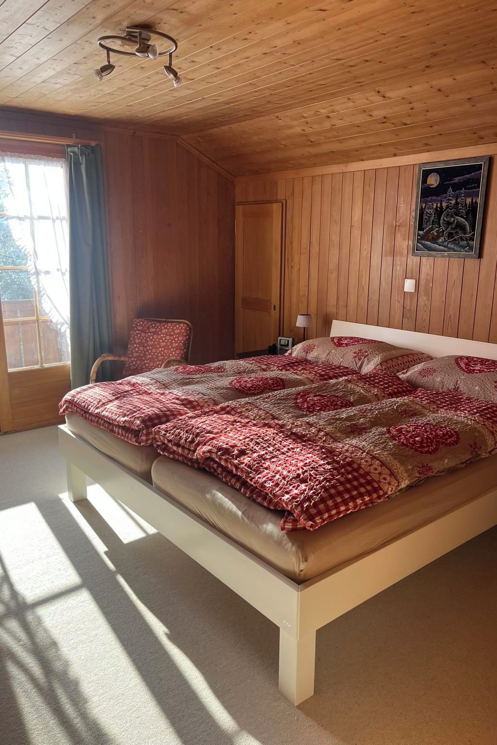 A bedroom with a wooden ceiling, a bed with a patterned blanket, a chair, a bedside table, and a window with curtains.