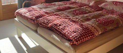 A bedroom with a wooden ceiling, a bed with a patterned blanket, a chair, a bedside table, and a window with curtains.