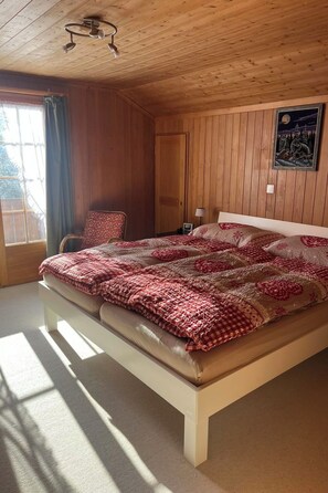 Ein Schlafzimmer mit Holzboden, einem Bett mit gemustertem Überwurf, einem Sessel, einem Nachttisch und einem Fenster mit Vorhängen.