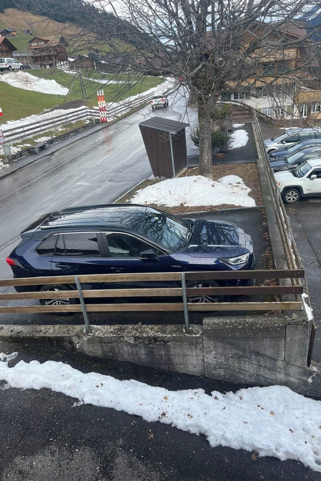 A blue car parked on a snowy roadside with a wooden fence and a small building in the background.
