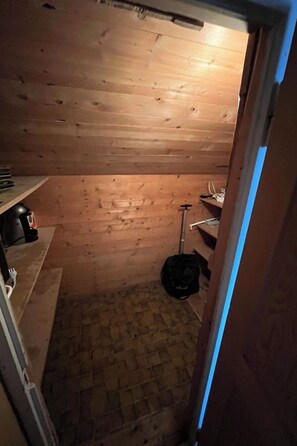 Ein kleiner, schummrig beleuchteter Raum mit Holzwänden und schräg verlaufender Decke. Ein einzelnes Regal mit einigen Gegenständen darauf, und eine schwarze Tasche hängt an der Wand.
